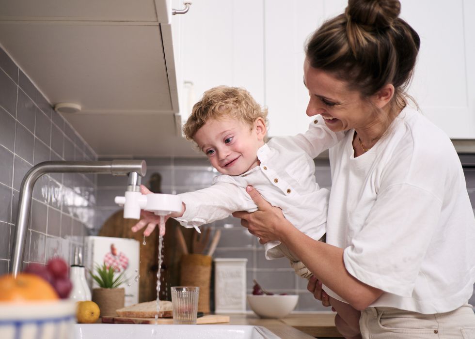 mamma håller sitt barn över diskbänken i köket med en rinnande vattenkran med vitt kranfilter från tappwater på, båda är glada och skrattar
