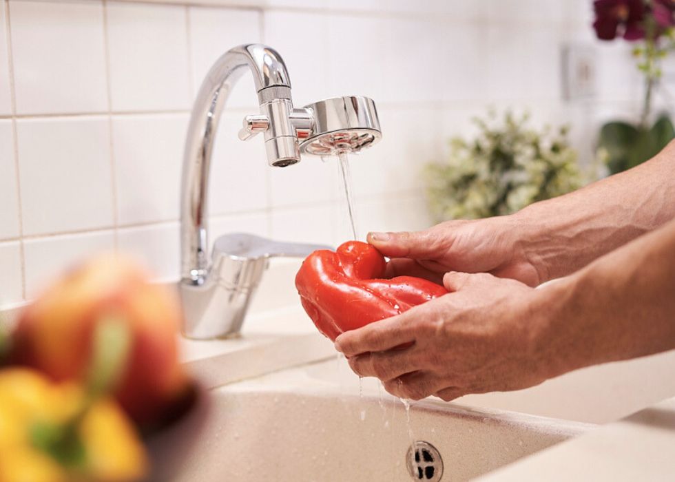 händer som håller i en röd paprika under en rinnande kran med kranfilter i krom på