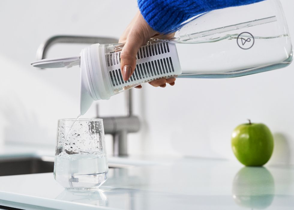 en arm med blå tröja håller i en vattenfilterkanna och häller upp renat vatten i glas, en rent fräscht kök med ett grön äpple i bakgrunden