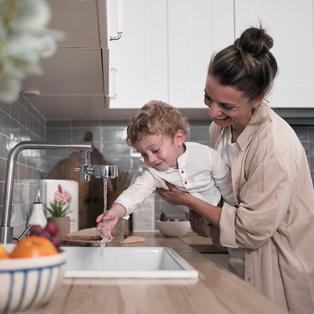 mamma håller barn som blöter handen i vatten från kranen, kranen har kranfilter
